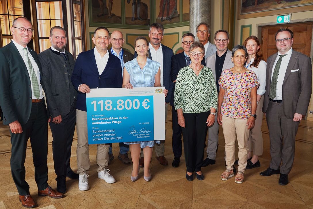 Scheckübergabe im Bayerischen Landtag: Die bayerische Staatsministerin für Gesundheit, Pflege und Prävention, Judith Gerlach, und der bpa-Landesvorsitzende Kai A. Kasri, umrahmt von (links) Bernhardt Seidenath, MdL CSU, und (hintere Reihe) Projektbeteiligten sowie Stefan Hahnemann und Joachim Görtz.